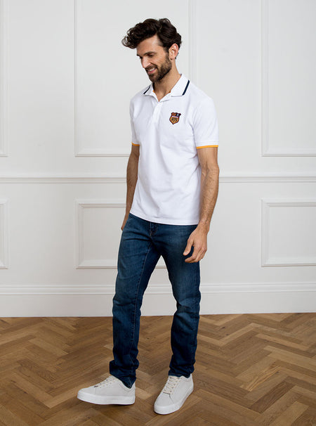 Man wearing white polo shirt in 100% cotton with Newmarket 875 crest embroidered on the front with navy sleeve trimming and gold collar trimming.